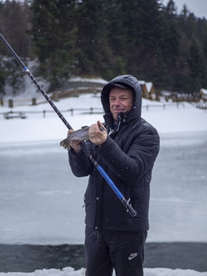 A person stands outdoors, holding a fishing rod and proudly displaying a fish in their hand. The surroundings are snowy, with a frozen body of water nearby and dense trees in the background. The person is dressed warmly in a black jacket with a hood.