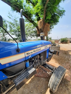 A blue tractor with 'New Holland' branding is parked near a large, leafy tree. The surroundings include a cobblestone ground and some farming equipment in the background. A haystack is positioned against the tree, and the sky is clear and sunny.