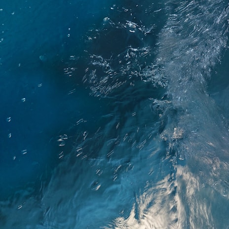 A detailed view of turbulent water, with visible ripples and waves creating a dynamic texture on the surface. The play of light on the water adds a sense of movement and depth.