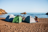 Three tents are pitched on a sandy beach beside the calm sea, with a couple of bicycles leaning against the tents. A person is organizing items near the tents, with rocky formations jutting out of the water in the background.