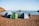Three tents are pitched on a sandy beach beside the calm sea, with a couple of bicycles leaning against the tents. A person is organizing items near the tents, with rocky formations jutting out of the water in the background.