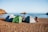 Three tents are pitched on a sandy beach beside the calm sea, with a couple of bicycles leaning against the tents. A person is organizing items near the tents, with rocky formations jutting out of the water in the background.