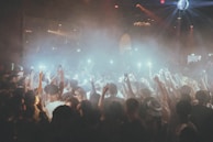 A crowded nightclub or concert scene with many people holding up their phones, capturing the moment with flashlights on. The atmosphere is energetic with fog and colorful lights, including beams and a disco ball above.