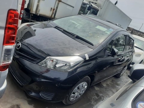 A black compact car is parked between other vehicles in a lot. The surface is wet, suggesting recent rain or washing. There are visible stickers on the windshield, and the paint appears reflective. Other parked vehicles, including a white van, are seen around it in the background.