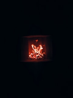A warm glow emanates from a small, controlled fire burning within a dark square fireplace. The flames flicker in shades of orange and red, providing a stark contrast to the surrounding darkness.