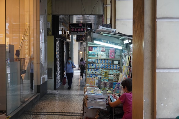 A narrow corridor in an urban setting features a small newsstand filled with newspapers and magazines. A woman wearing a pink shirt sits at the stand, appearing to be engaged in an activity. The pathway is lined with tiles and has a reflective glass display on one side, with people walking through the corridor.