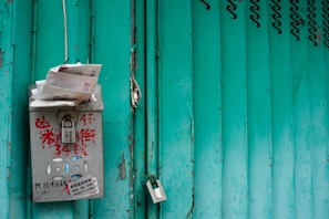 A gray metal mailbox with red text is mounted on a green gate. The mailbox is stuffed with letters and papers sticking out. The green gate has a series of vertical sections with noticeable wear and some locks hanging on it.