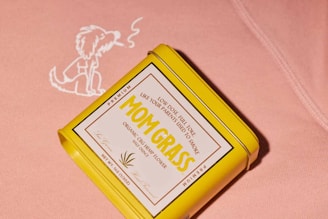 A yellow rectangular tin with a label reading 'MOM GRASS' is placed against a pink fabric background. The tin highlights it contains organic CBG hemp flower. Next to the tin on the fabric is an illustration of a dog exhaling smoke.