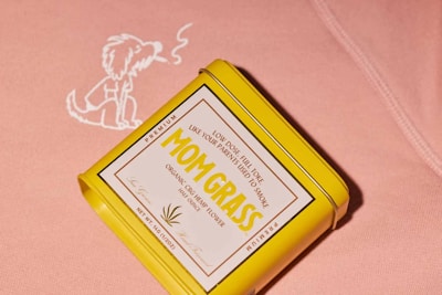 A yellow rectangular tin with a label reading 'MOM GRASS' is placed against a pink fabric background. The tin highlights it contains organic CBG hemp flower. Next to the tin on the fabric is an illustration of a dog exhaling smoke.
