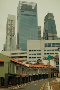 A street lined with traditional shophouses featuring colorful roofs and shutters is set against the backdrop of tall, modern skyscrapers. The architecture contrasts the historical and contemporary elements within an urban setting.