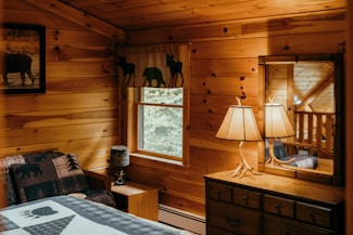 A cozy, rustic bedroom with wooden walls and warm lighting. There is a bed with a quilt featuring bear designs and a nearby nightstand adorned with a small lamp. A dresser with an antler-style lamp sits under a window, which is framed with curtains displaying animal silhouettes. Above the bed hangs a framed picture of an animal.