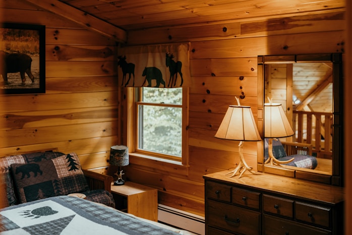 A cozy, rustic bedroom with wooden walls and warm lighting. There is a bed with a quilt featuring bear designs and a nearby nightstand adorned with a small lamp. A dresser with an antler-style lamp sits under a window, which is framed with curtains displaying animal silhouettes. Above the bed hangs a framed picture of an animal.