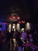 A dimly lit setting filled with people dancing energetically. The ceiling features several red lampshades that cast a warm glow over the room. The walls are dark with modern decor, and there is a disco ball reflecting light. The atmosphere appears lively and social.