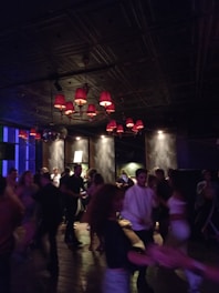 A dimly lit setting filled with people dancing energetically. The ceiling features several red lampshades that cast a warm glow over the room. The walls are dark with modern decor, and there is a disco ball reflecting light. The atmosphere appears lively and social.