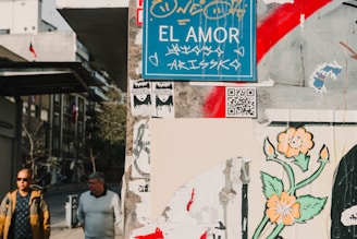 A street scene with a detailed blue sign reading 'EL AMOR' adorned with graffiti. It is surrounded by various posters and street art, including a floral design with orange flowers and a QR code. Two men are walking on the sidewalk in a city setting, with buildings and part of a flag in the background.