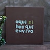 A brown canvas with text in soft green and turquoise colors reading 'aqui si hayqui enviva', placed against a gray wall. A small green plant is positioned on the left side.