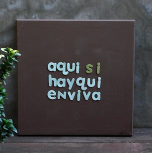 A brown canvas with text in soft green and turquoise colors reading 'aqui si hayqui enviva', placed against a gray wall. A small green plant is positioned on the left side.