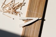 A collection of cotton swabs lies scattered next to a wooden sushi rolling mat. A white plastic tool with a textured grip rests on top of the mat, casting a shadow. The surface is well-lit, highlighting the texture and contrast between the objects.