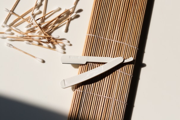 A collection of cotton swabs lies scattered next to a wooden sushi rolling mat. A white plastic tool with a textured grip rests on top of the mat, casting a shadow. The surface is well-lit, highlighting the texture and contrast between the objects.