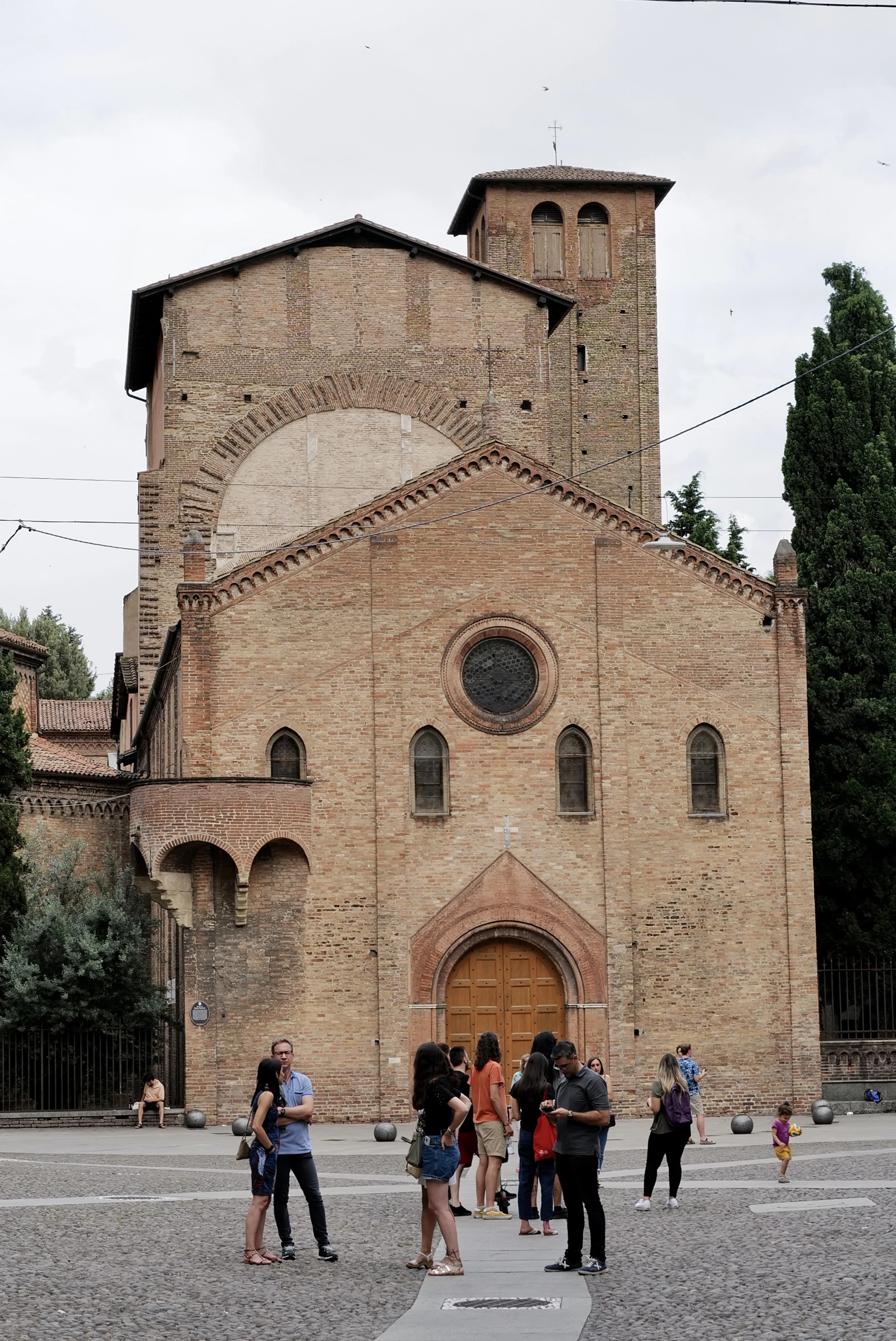 The brick Romanesque facade of the Basilica di Santo Stefano in Bologna, with visitors gathered in the piazza outside.