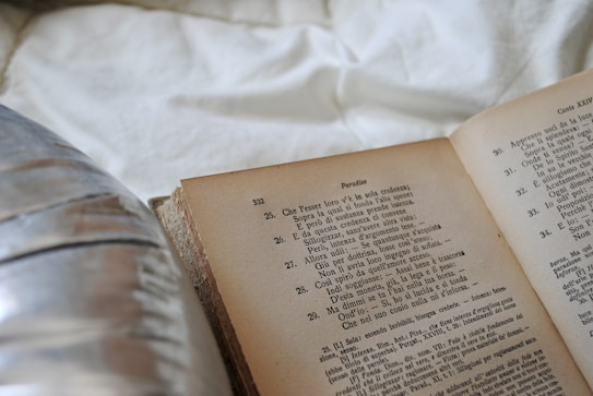 An open book with text in Italian is resting on a white fabric surface. To the left, a blurred reflective surface, possibly a metal object, is partially visible.