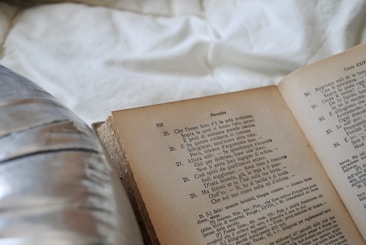 An open book with text in Italian is resting on a white fabric surface. To the left, a blurred reflective surface, possibly a metal object, is partially visible.