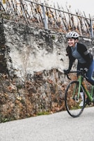 A person wearing a helmet and cycling gear is riding a green bicycle along a road. The background features a rugged stone wall with sparse vegetation and a row of leafless vines supported by stakes.