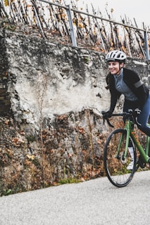 A person wearing a helmet and cycling gear is riding a green bicycle along a road. The background features a rugged stone wall with sparse vegetation and a row of leafless vines supported by stakes.