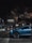 A blue luxury car is parked on a cobbled street at night in front of a restaurant named Coco's Kitchen, with warm lights glowing from inside. Several people can be seen walking near the entrance, and trees partially obscure the top part of the building.