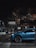 A blue luxury car is parked on a cobbled street at night in front of a restaurant named Coco's Kitchen, with warm lights glowing from inside. Several people can be seen walking near the entrance, and trees partially obscure the top part of the building.