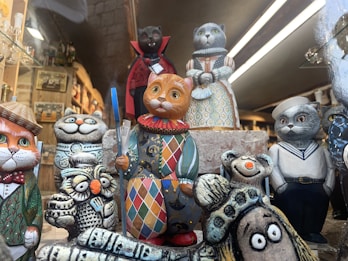 A collection of intricately painted ceramic cat figurines arranged together. Each cat is dressed in colorful, ornate costumes, from a sailor uniform to a noblewoman's dress, and one holding a book. The scene also includes additional animal figurines like owls with similar artistic styles. The setting appears to be a shop or display area with shelves filled with various items in the background.