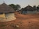 Traditional round thatched-roof huts are situated in a rural village setting. The huts have earthen walls painted with patterns, and there is sparse vegetation in the surrounding red soil. A few small trees provide some greenery amid the dry landscape.
