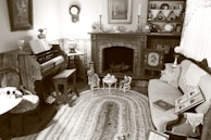 A vintage living room with an antique organ on the left, a decorative fireplace mantel adorned with candlesticks and ornate ceramics, and a cozy seating area with a patterned sofa. A woven rug covers the floor, and shelves hold books and small collectibles. The atmosphere is quaint and nostalgic, with lace doilies and framed pictures accentuating the old-fashioned charm.