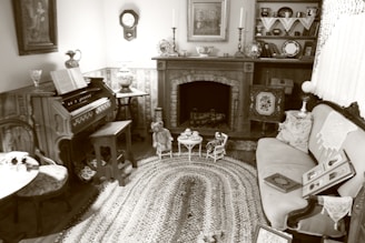 A vintage living room with an antique organ on the left, a decorative fireplace mantel adorned with candlesticks and ornate ceramics, and a cozy seating area with a patterned sofa. A woven rug covers the floor, and shelves hold books and small collectibles. The atmosphere is quaint and nostalgic, with lace doilies and framed pictures accentuating the old-fashioned charm.