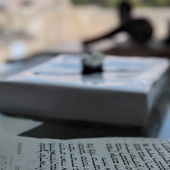 A close-up of a text with Hebrew script, partially in focus, on a blurred background that includes a window view, suggesting an indoor setting. The surface of the paper is slightly glossy.