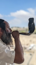 A man with a beard is blowing a shofar, an ancient musical horn typically associated with Jewish religious practices. He is wearing a white shirt and a black cap. The background is slightly blurred, showing an outdoor setting with a blue sky and some scattered clouds.