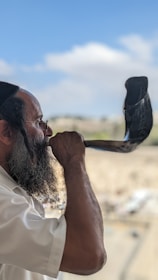 A man with a beard is blowing a shofar, an ancient musical horn typically associated with Jewish religious practices. He is wearing a white shirt and a black cap. The background is slightly blurred, showing an outdoor setting with a blue sky and some scattered clouds.