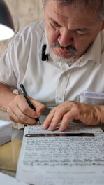 A person with a beard is sitting at a desk, focused on writing on a sheet of paper with a pen. The paper is filled with handwritten text. The person is wearing a white shirt and appears to be concentrating on the task. A ruler is placed on the paper.