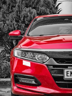 A vibrant red car is prominently displayed, hinting at a sporty or sleek look. The car's design features sharp lines and a modern headlight design. It is parked near lush, dark green foliage which contrasts dramatically with the bright color of the car.