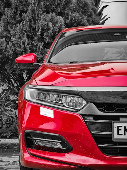 A vibrant red car is prominently displayed, hinting at a sporty or sleek look. The car's design features sharp lines and a modern headlight design. It is parked near lush, dark green foliage which contrasts dramatically with the bright color of the car.