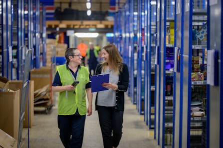 Two individuals wearing work attire are walking through a warehouse aisle, engaged in conversation. The shelves are lined with various products and items, and both people hold clipboards. The setting is well-lit, highlighting the organized shelves.