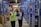 Two individuals wearing work attire are walking through a warehouse aisle, engaged in conversation. The shelves are lined with various products and items, and both people hold clipboards. The setting is well-lit, highlighting the organized shelves.