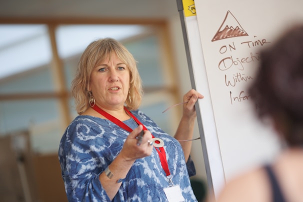 A woman with short blonde hair, wearing a blue patterned shirt and a red lanyard, is standing in front of a whiteboard. She appears to be teaching or presenting, holding a pointer in her right hand. The whiteboard contains a drawing of a mountain and several handwritten points, including the words 'ON Time' and 'Objectives'. There is a blurred figure in the foreground listening attentively.