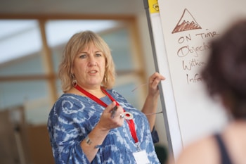 A woman with short blonde hair, wearing a blue patterned shirt and a red lanyard, is standing in front of a whiteboard. She appears to be teaching or presenting, holding a pointer in her right hand. The whiteboard contains a drawing of a mountain and several handwritten points, including the words 'ON Time' and 'Objectives'. There is a blurred figure in the foreground listening attentively.