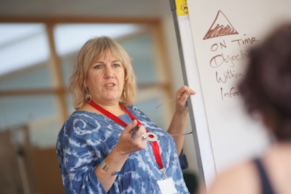 A woman with short blonde hair, wearing a blue patterned shirt and a red lanyard, is standing in front of a whiteboard. She appears to be teaching or presenting, holding a pointer in her right hand. The whiteboard contains a drawing of a mountain and several handwritten points, including the words 'ON Time' and 'Objectives'. There is a blurred figure in the foreground listening attentively.