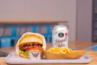 A juicy burger topped with lettuce, tomato, and sauce is neatly wrapped in branded paper on a rectangular tray. Beside it, a portion of crispy golden fries is served in a brown paper holder, and a can of Hopped Cider is placed next to the food. The setting includes blurred blue chairs and wooden tables, indicating a casual dining environment.