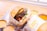 A gourmet burger is wrapped in paper labeled 'Savouri Eatery' and placed on a tray. The burger consists of a thick beef patty, lettuce, tomato, sauce, and melted cheese. Surrounding the burger are small metal cups filled with different dipping sauces.