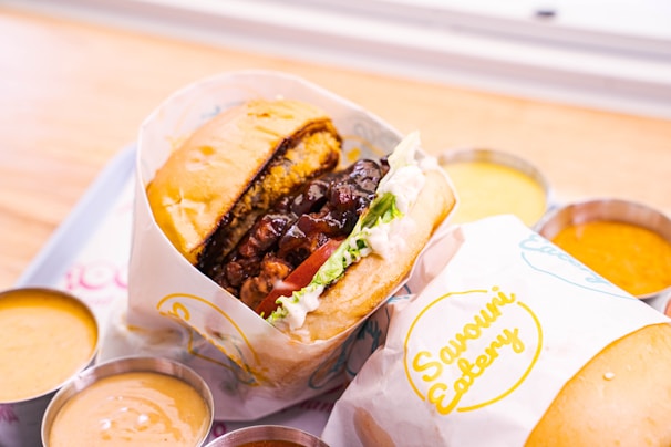A gourmet burger is wrapped in paper labeled 'Savouri Eatery' and placed on a tray. The burger consists of a thick beef patty, lettuce, tomato, sauce, and melted cheese. Surrounding the burger are small metal cups filled with different dipping sauces.