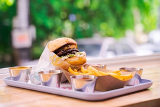 A juicy hamburger wrapped partially in branded paper is placed on a tray alongside a serving of golden, crispy fries. Small metal cups, presumably for sauces, are arranged around the food. The background features a soft-focus view of green foliage outside, suggesting an outdoor or well-lit indoor setting.