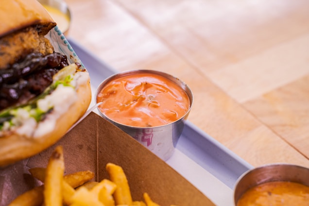 A close-up of a burger with melted cheese and sauce, accompanied by a metal cup filled with orange dipping sauce. Crinkle-cut fries are partially visible in a cardboard container, all set on a wooden table.